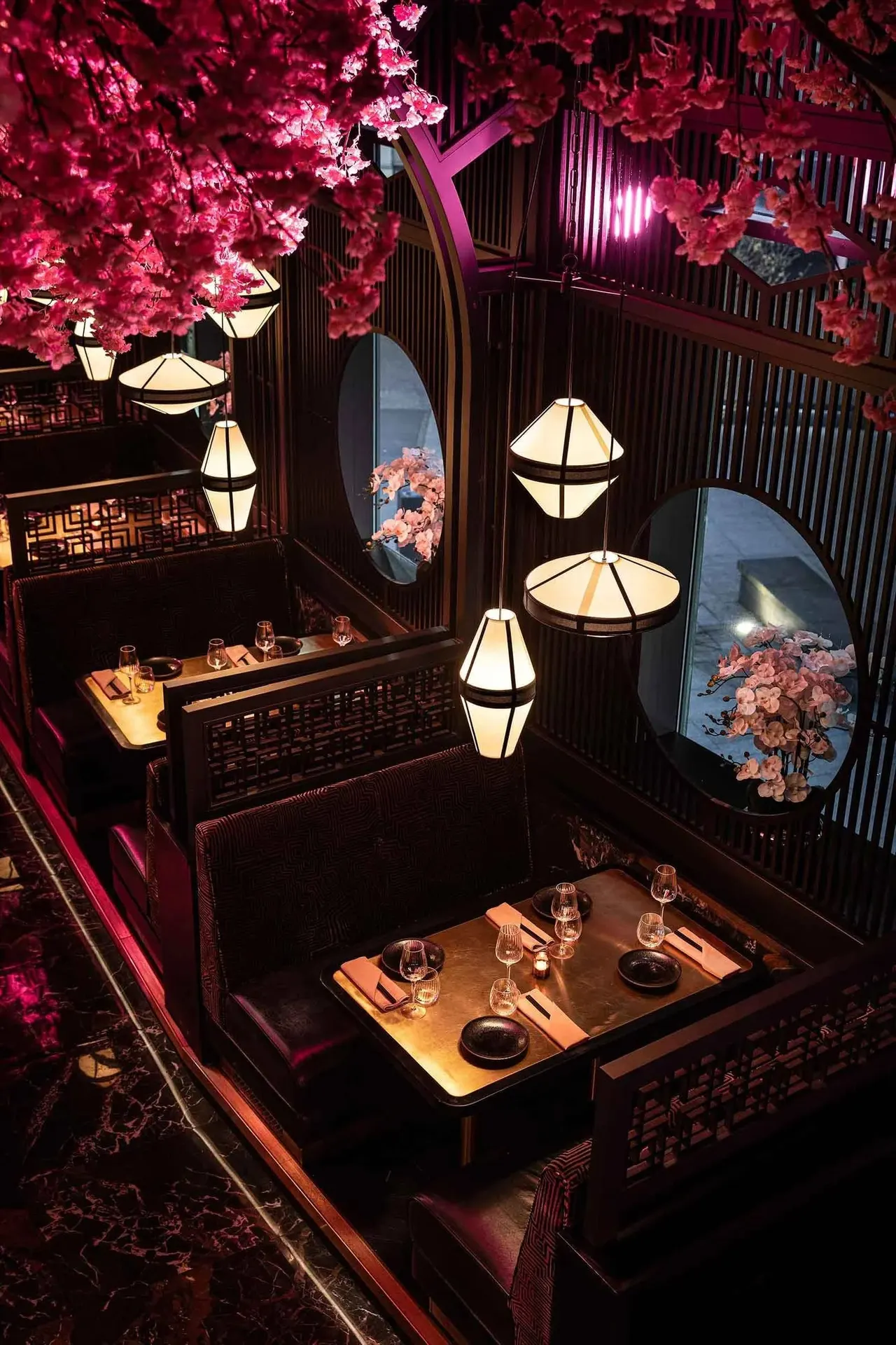 Overhead view of an elegant dining booth with set tables, illuminated lanterns and vibrant pink flowering branches in an Asian-inspired restaurant interior.