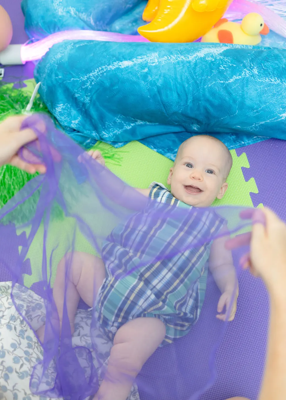 Baby lying on colourful play mat surrounded by bright toys and sensory play equipment.