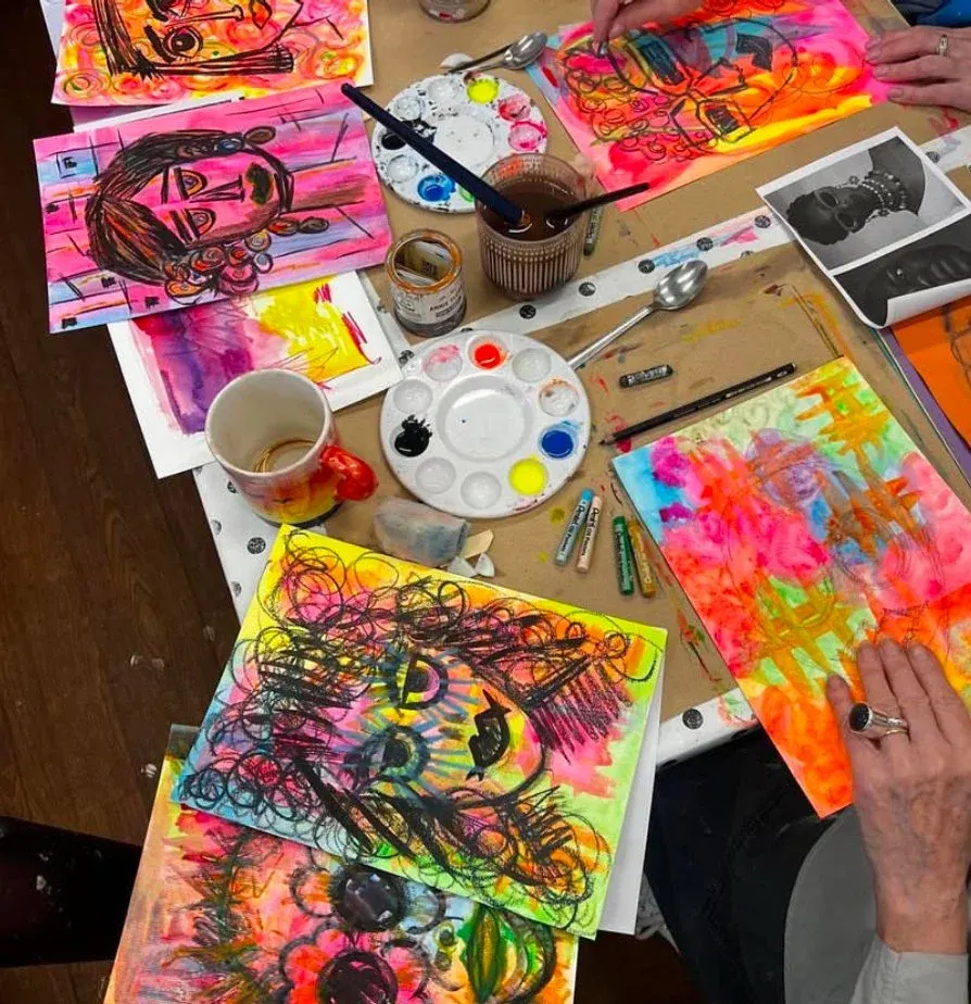 People creating colourful painted artwork at a table surrounded by paintbrushes, palettes, and finished vibrant designs.