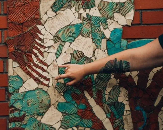 Person pointing at a close-up detail of ornate street art or mural featuring geometric patterns in red, green, turquoise and cream.
