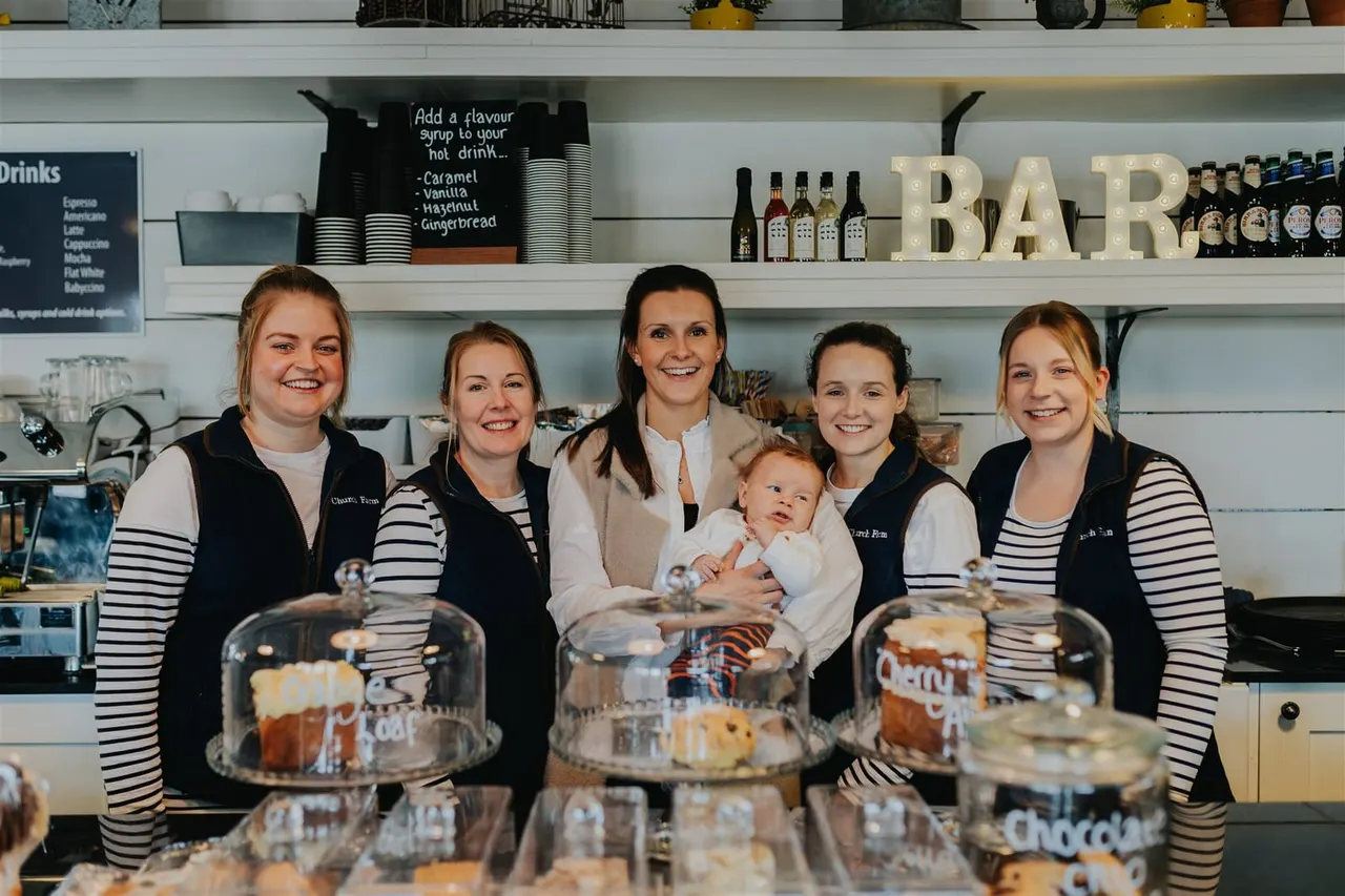 Six smiling café staff members and a small child stand behind the counter at a tea rooms, with shelved drinks, bottles, and BAR signage visible behind them.