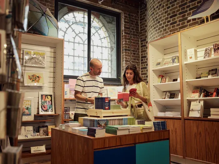 Two people browse and handle books at a counter inside a brick-walled bookshop with large arched windows and shelving displays.