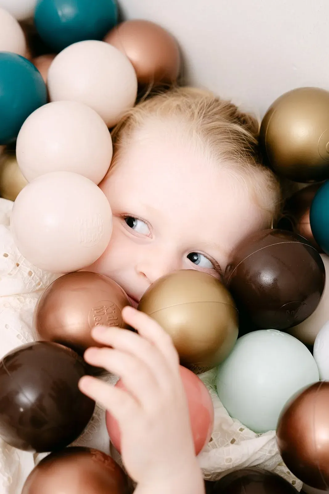 A young child playing in a ball pit filled with cream, bronze, teal, and brown balls.