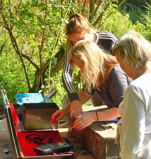 Three people examining contents of a red container outdoors amongst green foliage and dappled sunlight.
