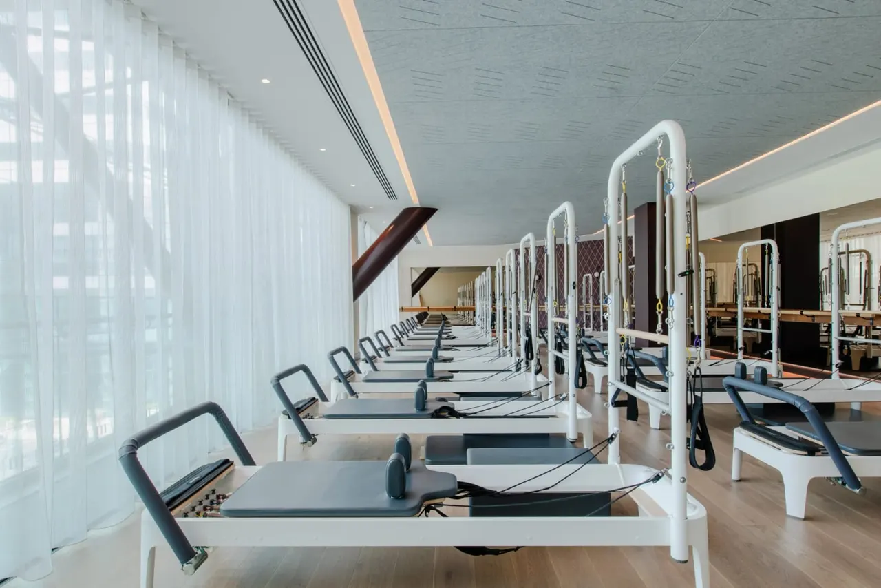 A modern pilates studio with rows of reformer machines lined up along a bright, naturally lit space with floor-to-ceiling windows.