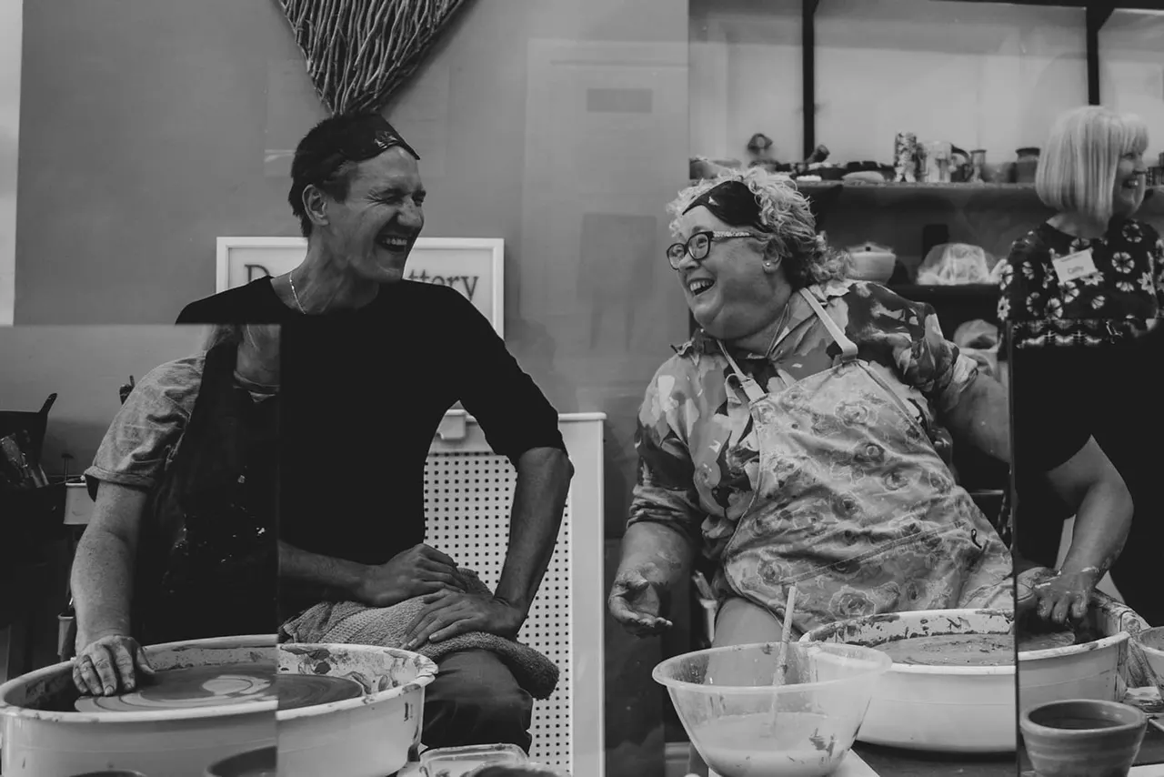 Two people laughing whilst preparing pottery, with clay bowls and materials on a work table in a studio.