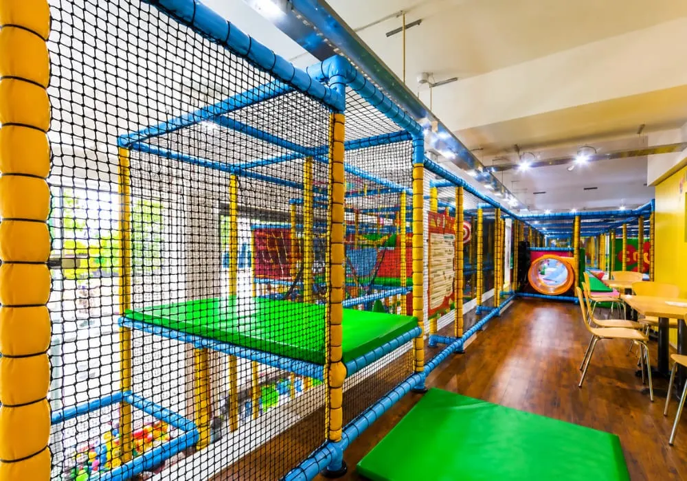 Indoor children's play area with blue and yellow framed net structures containing green padded surfaces and various coloured play equipment.