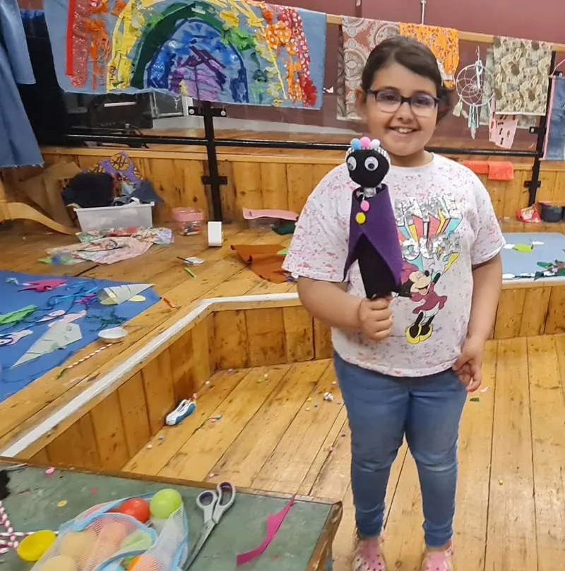 Child holding a handmade puppet in an art studio with colourful artwork displayed on walls and wooden tables.