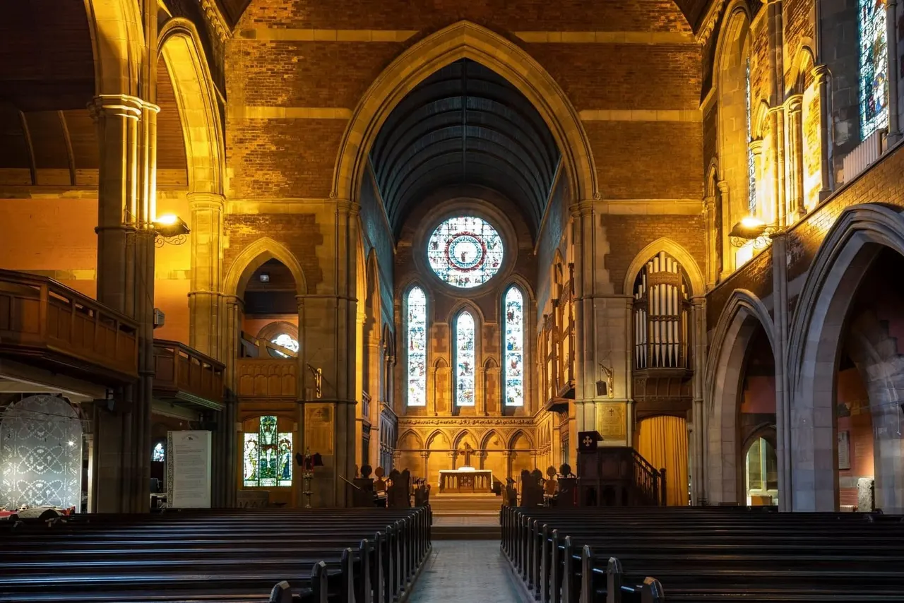 Interior of a Gothic church with vaulted ceilings, ornate stonework, illuminated archways, stained glass windows and a large clock face above the altar.