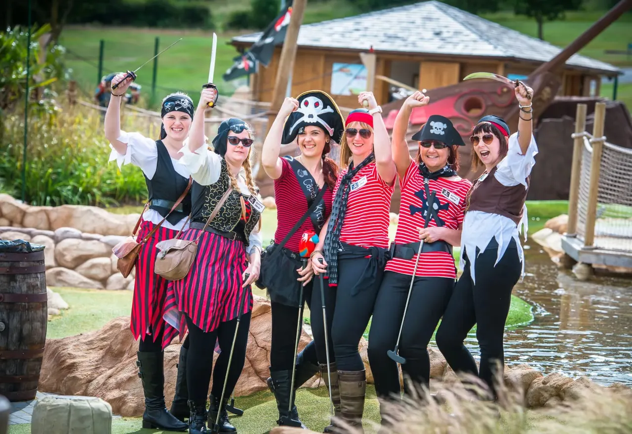 A group of six people dressed in pirate costumes pose together holding fishing rods at a mini golf venue, with a wooden lodge and water feature visible behind them.