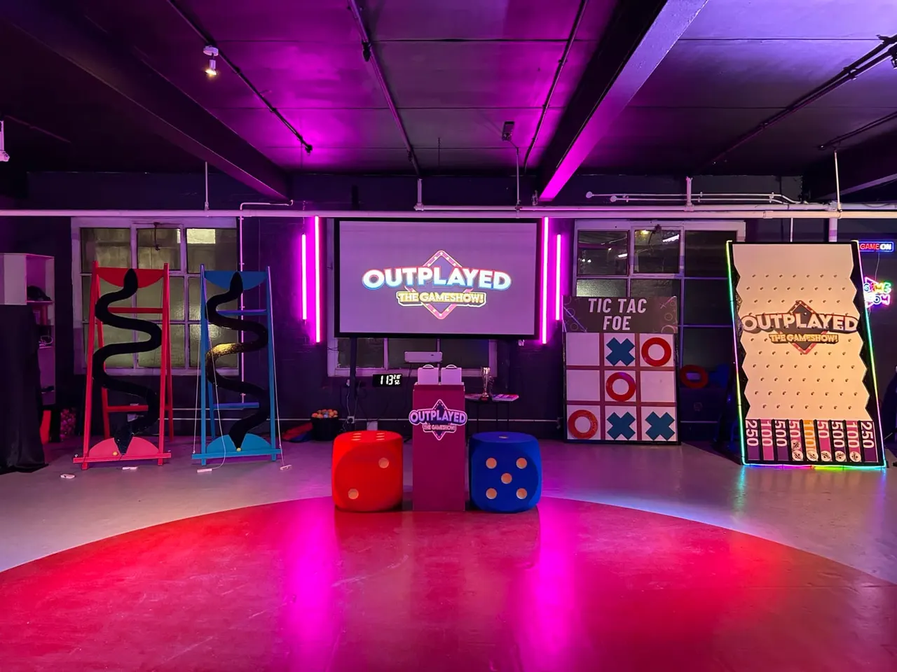 Indoor games venue with neon-lit interactive game stations, oversized dice, and climbing activities under purple lighting.