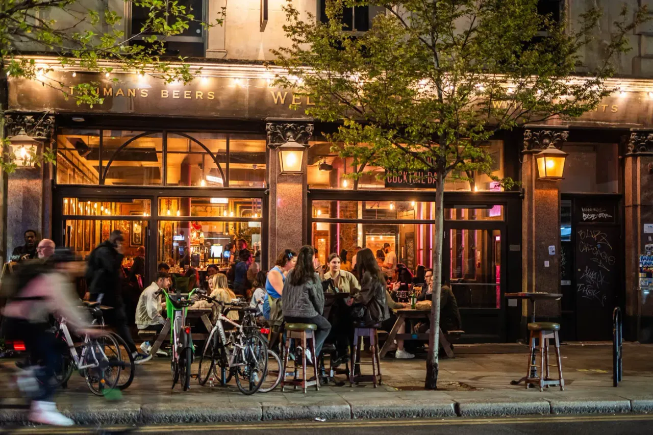 Evening street scene with people standing and sitting outside an illuminated bar, some with bicycles parked nearby.