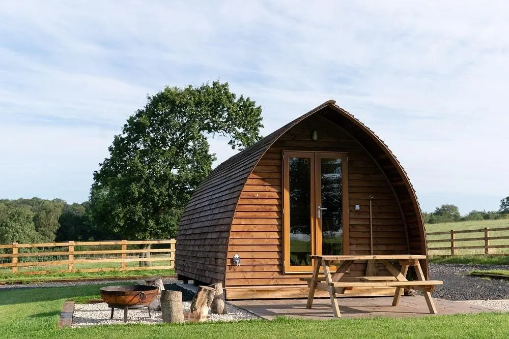 Glamping pod with arched roof and wooden seating area in a rural pasture setting with fields and trees beyond.