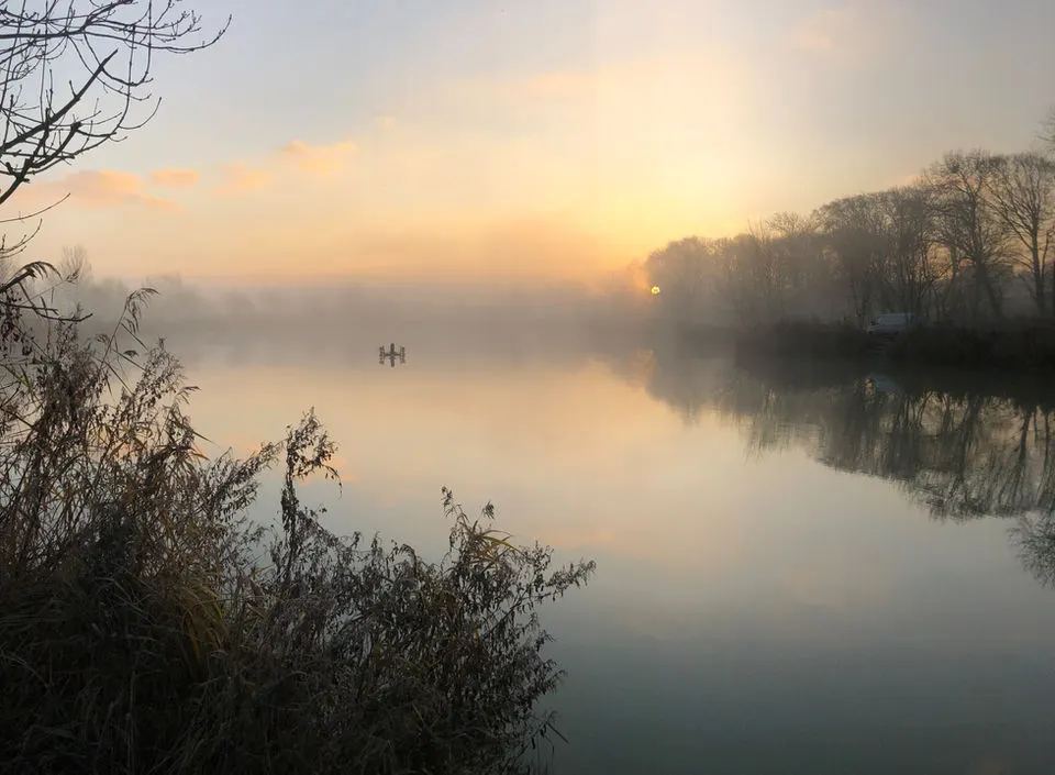 Misty sunrise over a calm river with bare winter trees and a small boat in the distance.