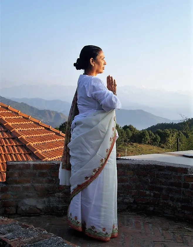 Woman in white traditional dress practising yoga on a rooftop terrace with mountains in the background.