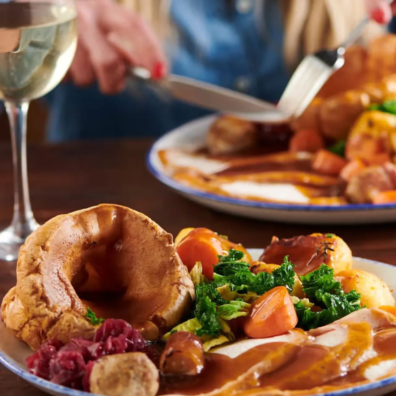 People dining with roast dinner plates featuring Yorkshire pudding, vegetables and gravy, with wine glasses on the table.