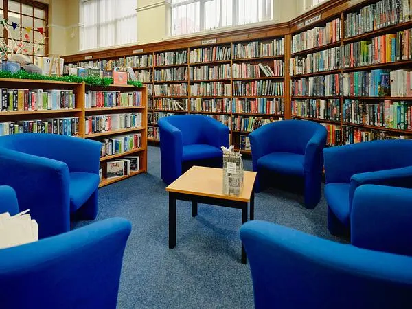 Bright library reading area with blue armchairs arranged around a low table, surrounded by floor-to-ceiling bookshelves.