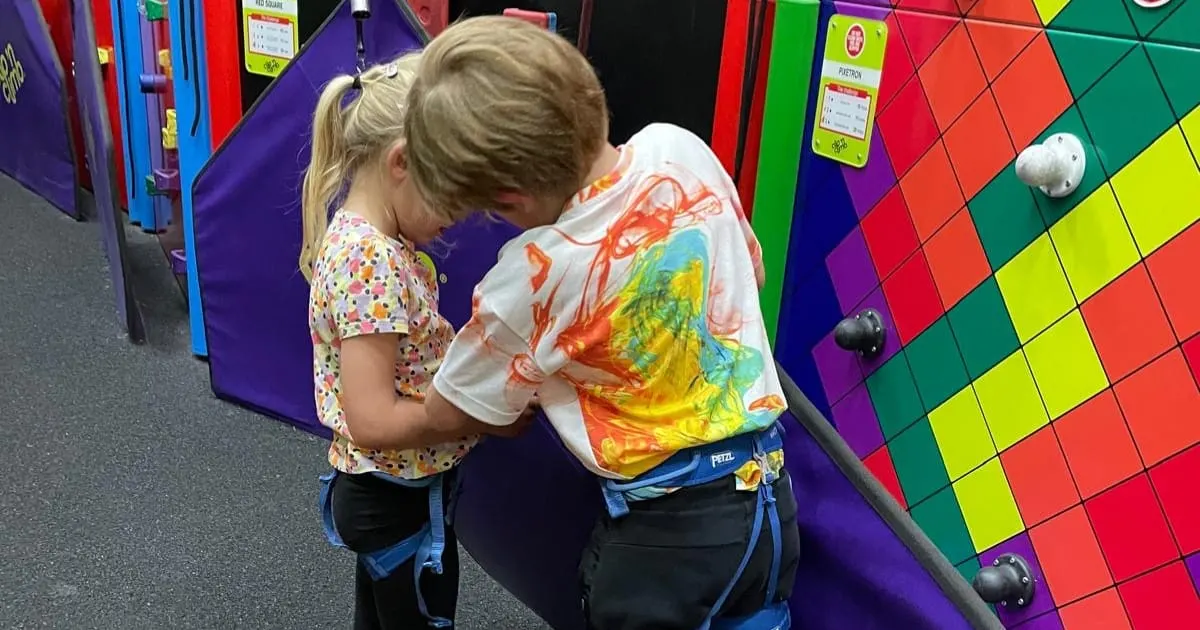 Two children climbing an indoor coloured wall with safety harnesses at a climbing centre.