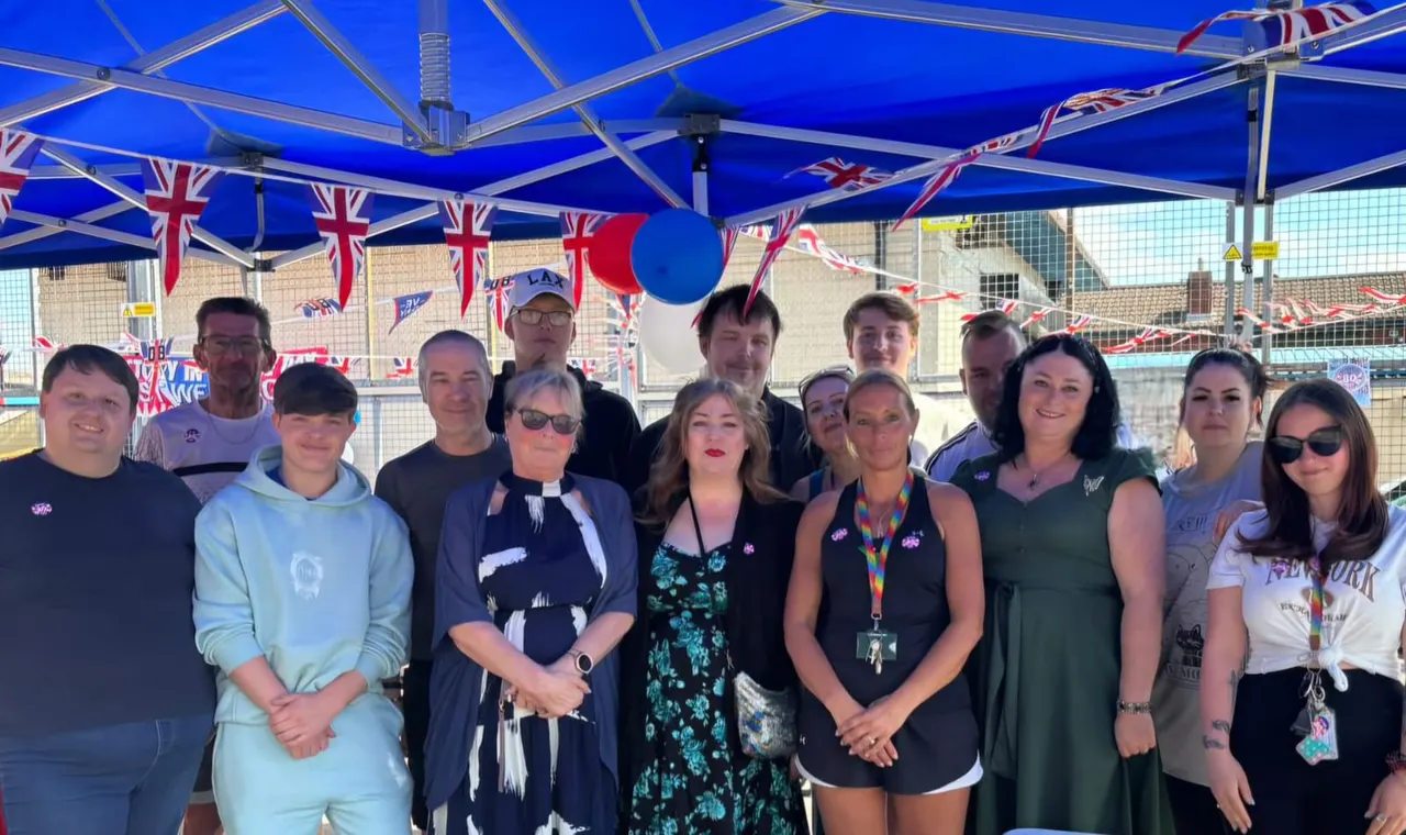 Group of people posing together under a blue marquee decorated with Union Jack bunting and balloons at an outdoor event.