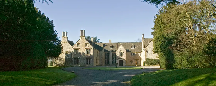 Stone country manor house with manicured lawns and tree-lined drive under clear blue sky.