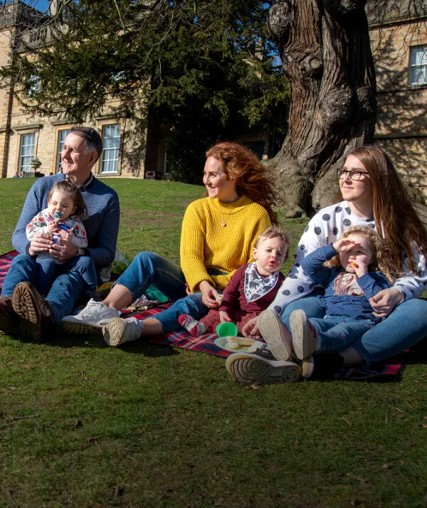 Two adults and four young children sitting on grass with a picnic in front of a historic stone house and large tree.