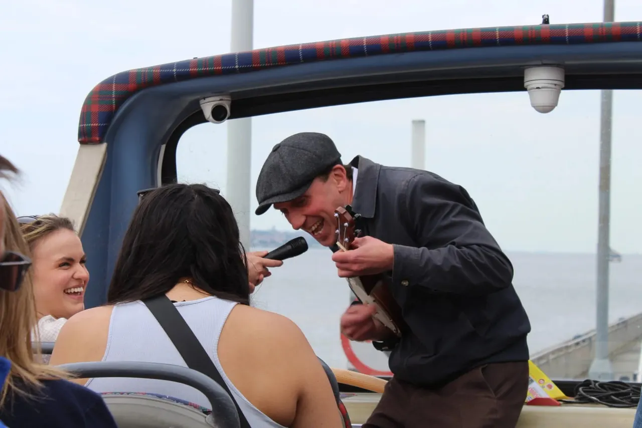 A man with a microphone entertains laughing passengers on an open-top tour bus overlooking a waterfront.