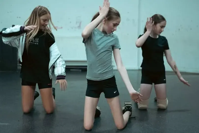 Three young children kneeling on a dark floor during a dance or movement class.