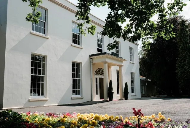 White neoclassical building with columned entrance, ivy-covered walls, and colourful flower beds in foreground.