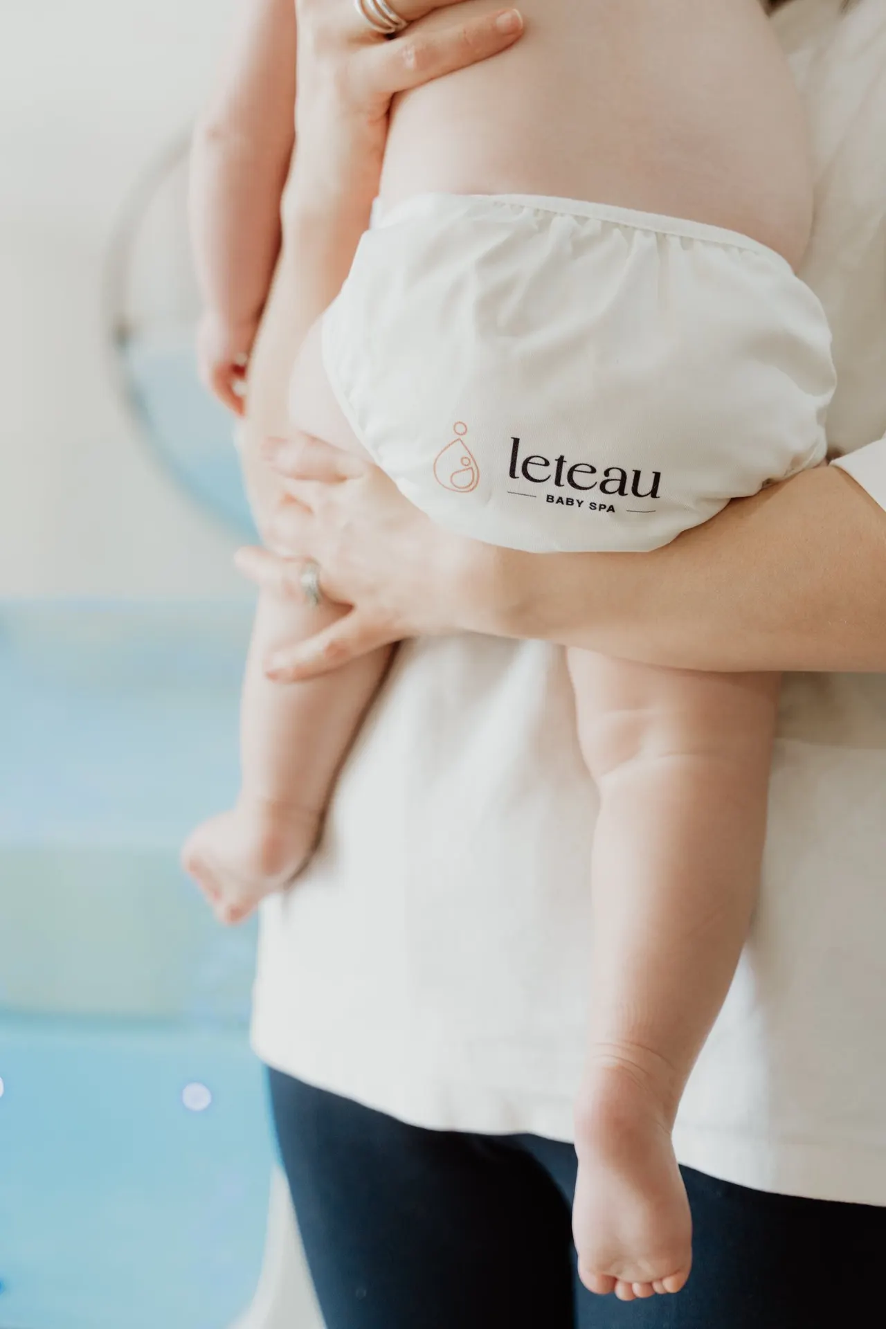 Adult holding infant wearing branded nappy near water, poolside setting.