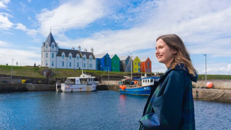 Woman standing at harbour looking towards colourful beachside buildings and moored fishing boats.