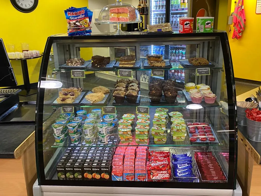 A curved glass display counter filled with cakes, brownies, confectionery and packaged snacks inside a bright yellow café.