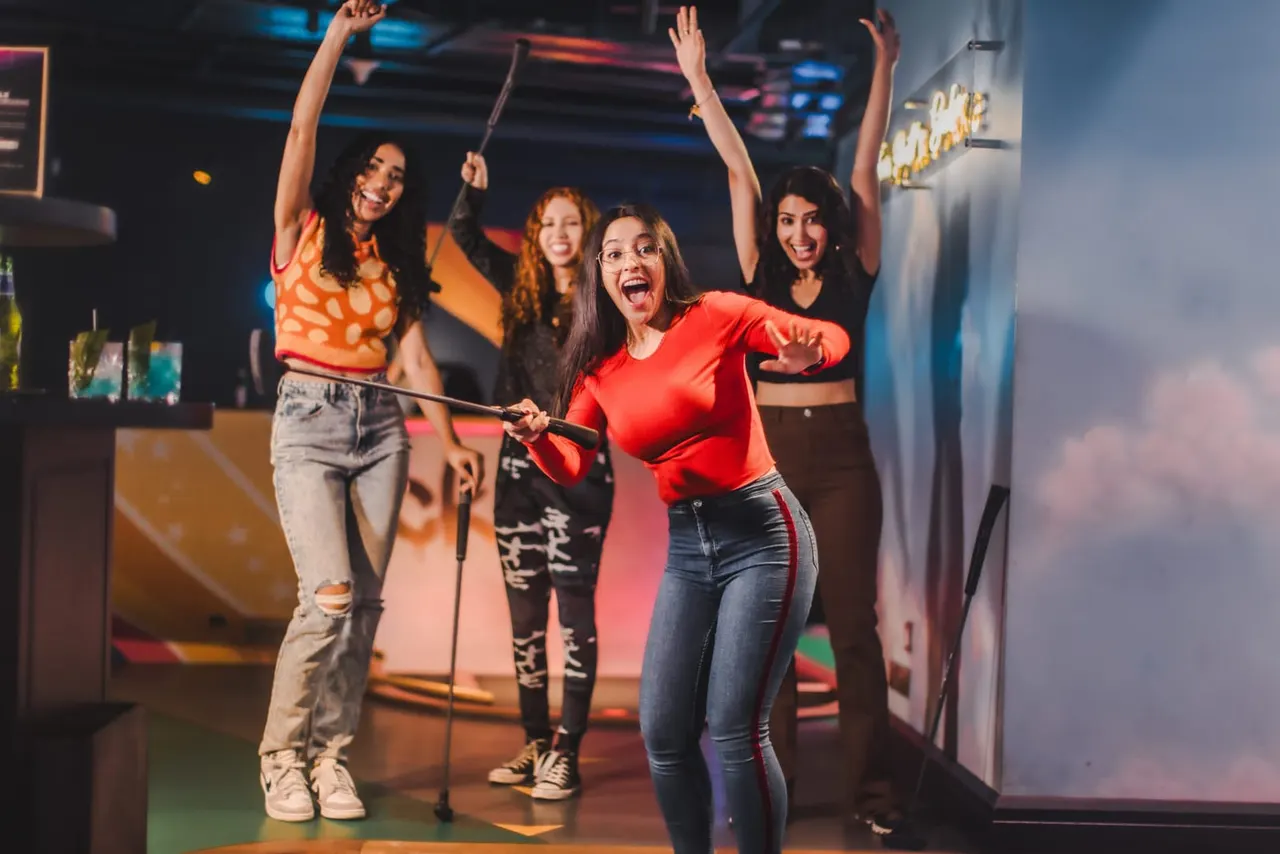 A group of five women singing and dancing together on a karaoke stage with colourful neon lighting and holding microphones.