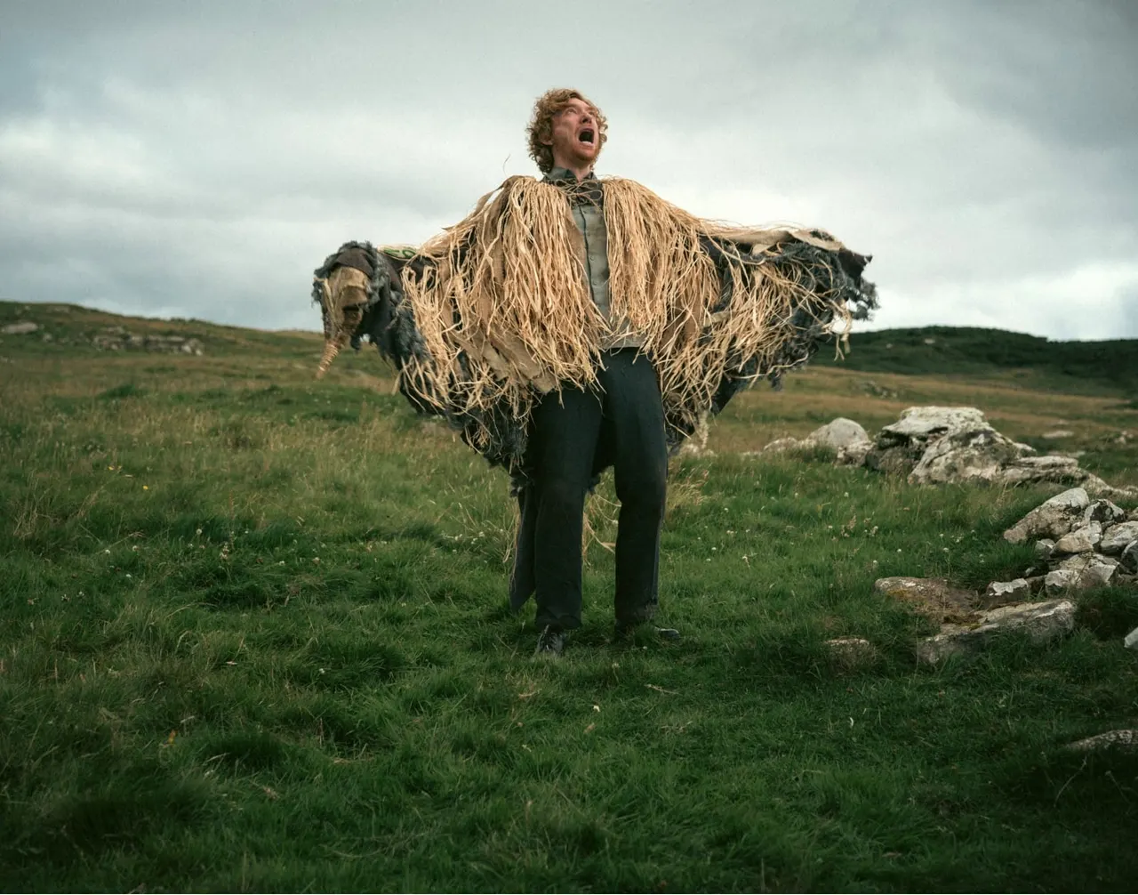 Man wearing a straw cloak with animal heads stands in open moorland, looking upward dramatically.