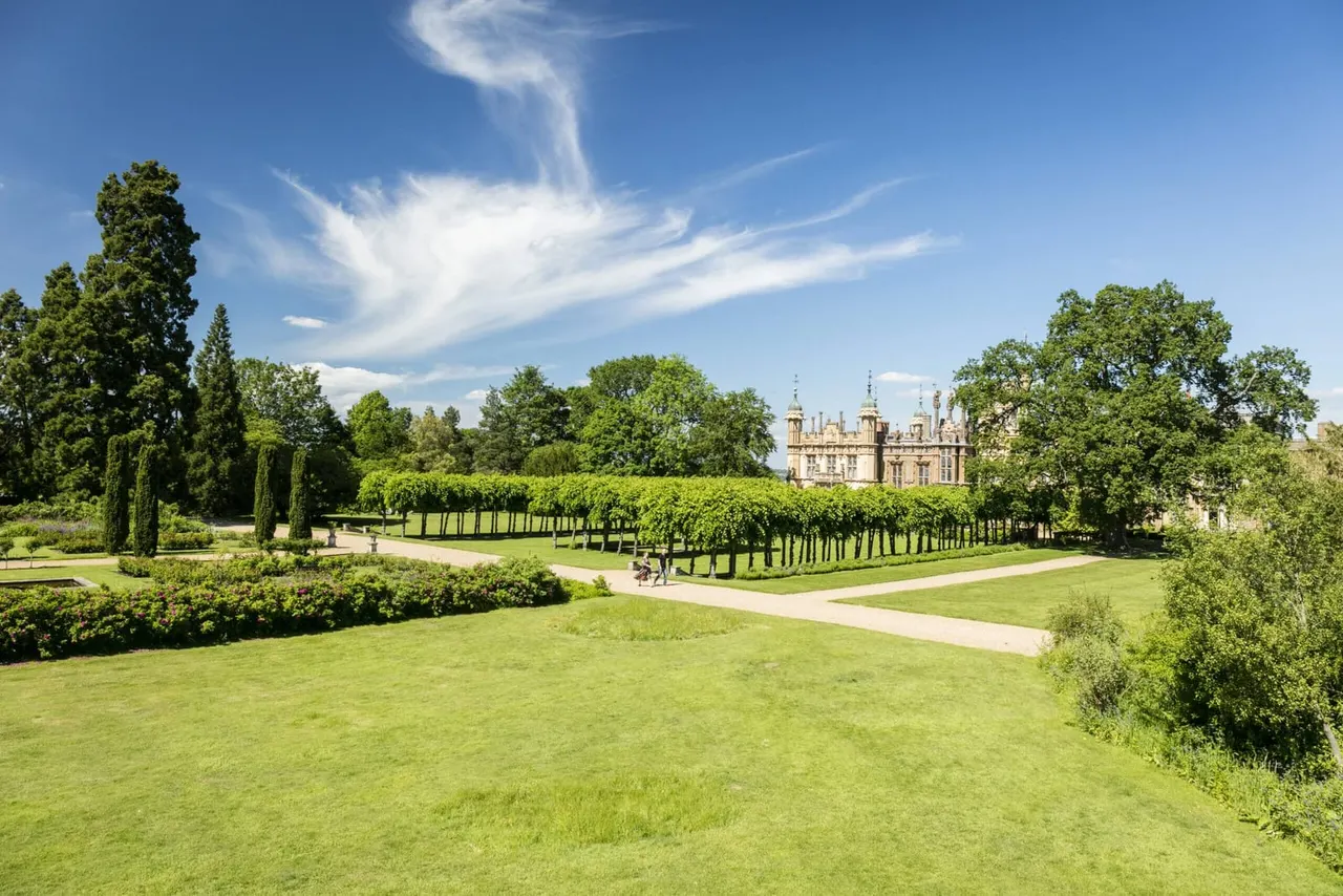Expansive manicured gardens with tree-lined pathways leading toward an ornate stone manor house on a sunny day.