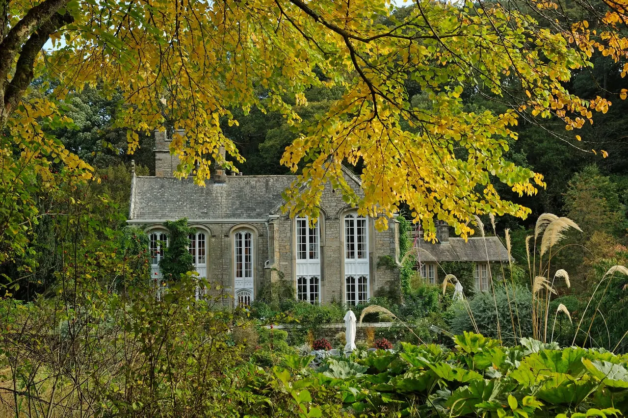 Stone chapel with ivy-covered walls framed by golden autumn foliage and surrounded by planted gardens.