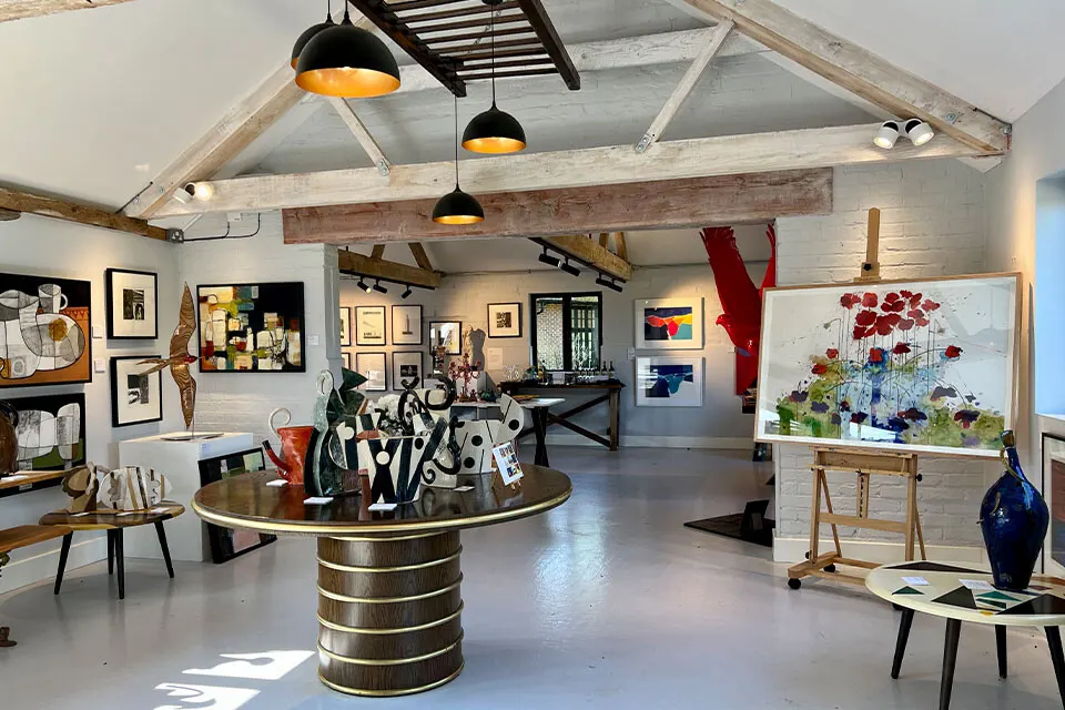 Interior of an art gallery with exposed wooden beams, pendant lights, and various artworks displayed on white walls and easels.