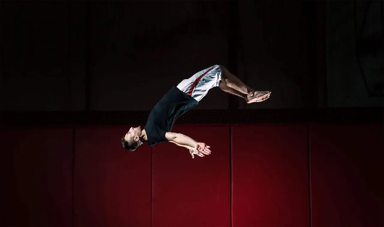 Acrobat performing a backflip in mid-air against a red wall at an indoor trampoline park.