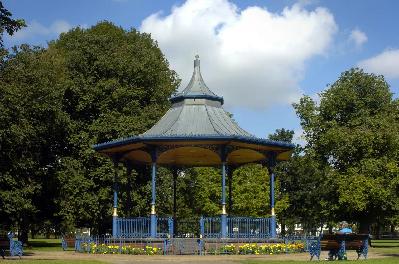 A blue and yellow Victorian bandstand with a pointed roof surrounded by mature trees and yellow flowers in a park setting.