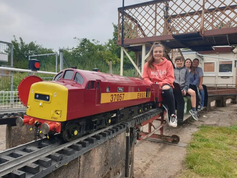 Children and adults sitting in a red and yellow miniature railway carriage at an outdoor heritage railway venue.