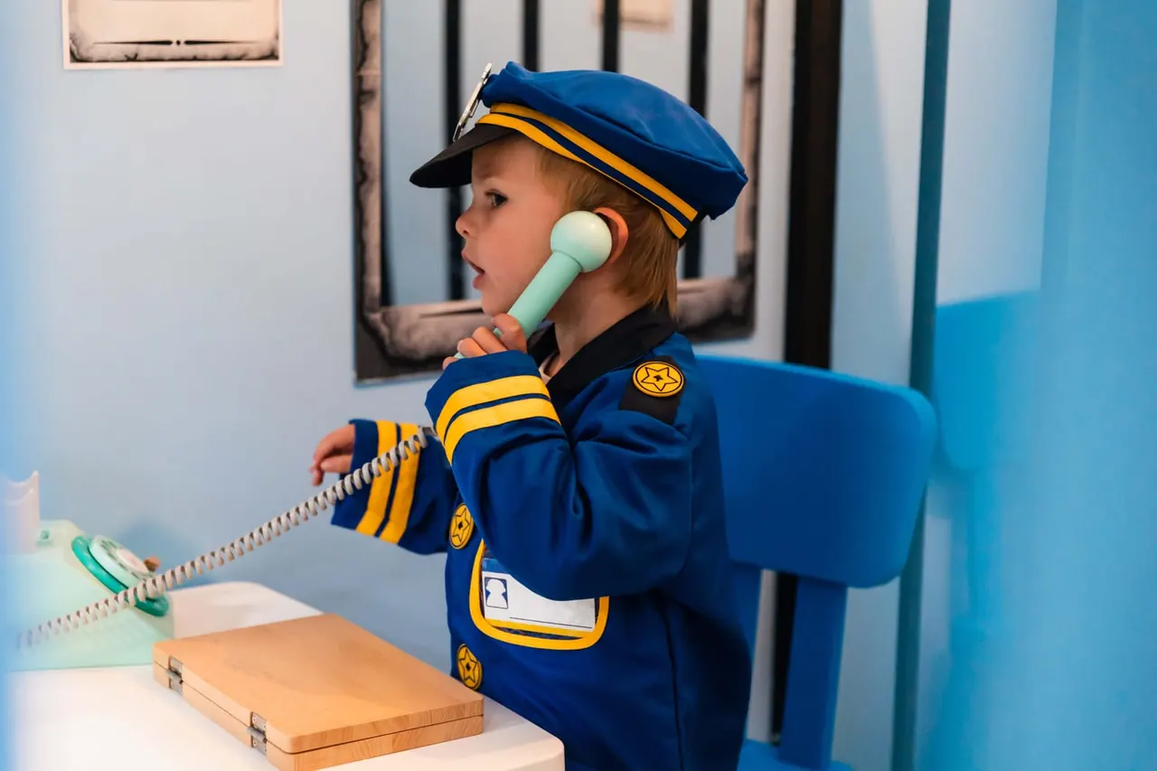 Young child in police officer costume speaking into toy telephone at play village.