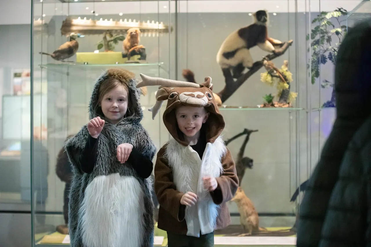 Two young children in animal costumes pose in front of a museum display of taxidermied animals behind glass.