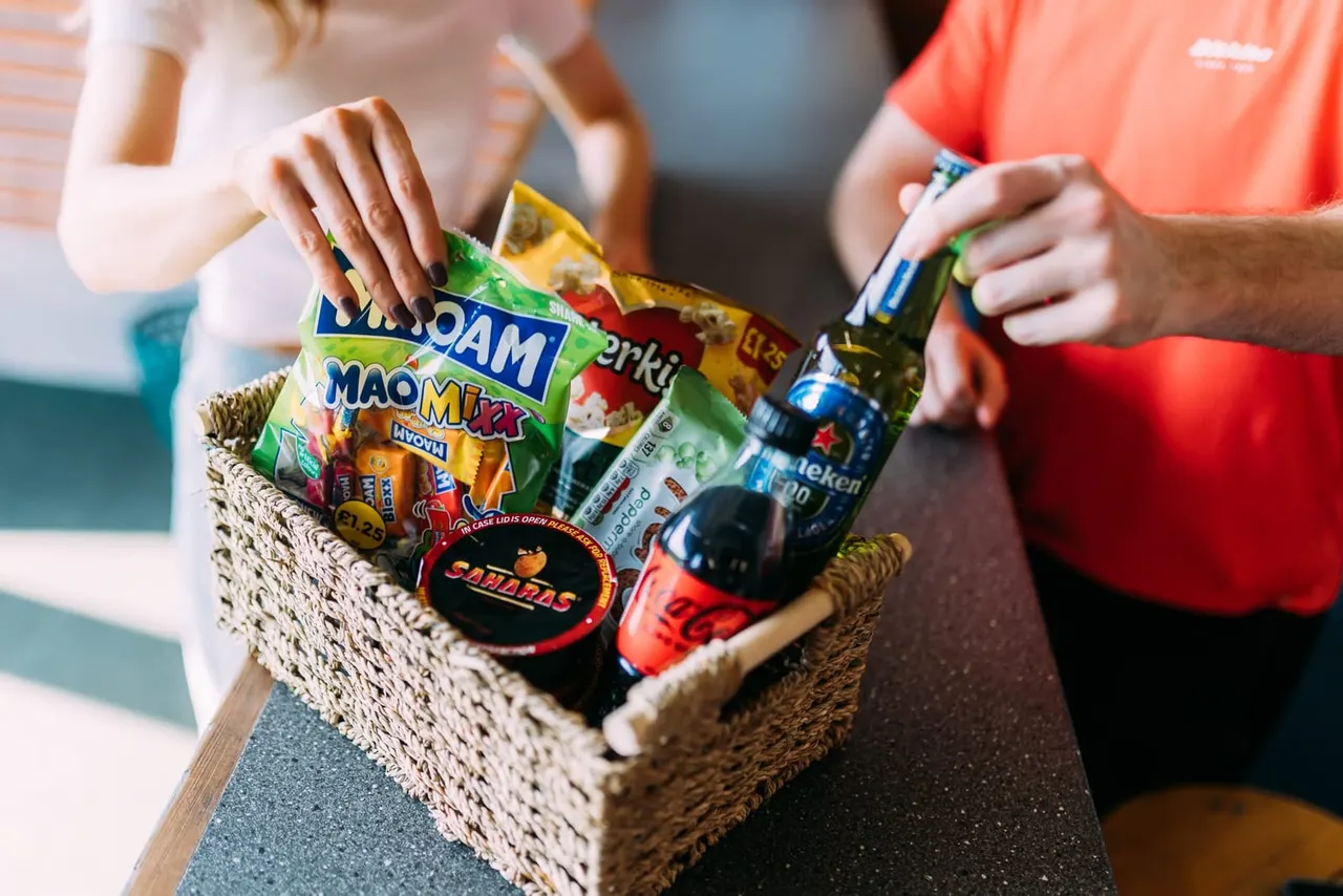 Two people holding a woven basket filled with snacks, beverages and branded items.