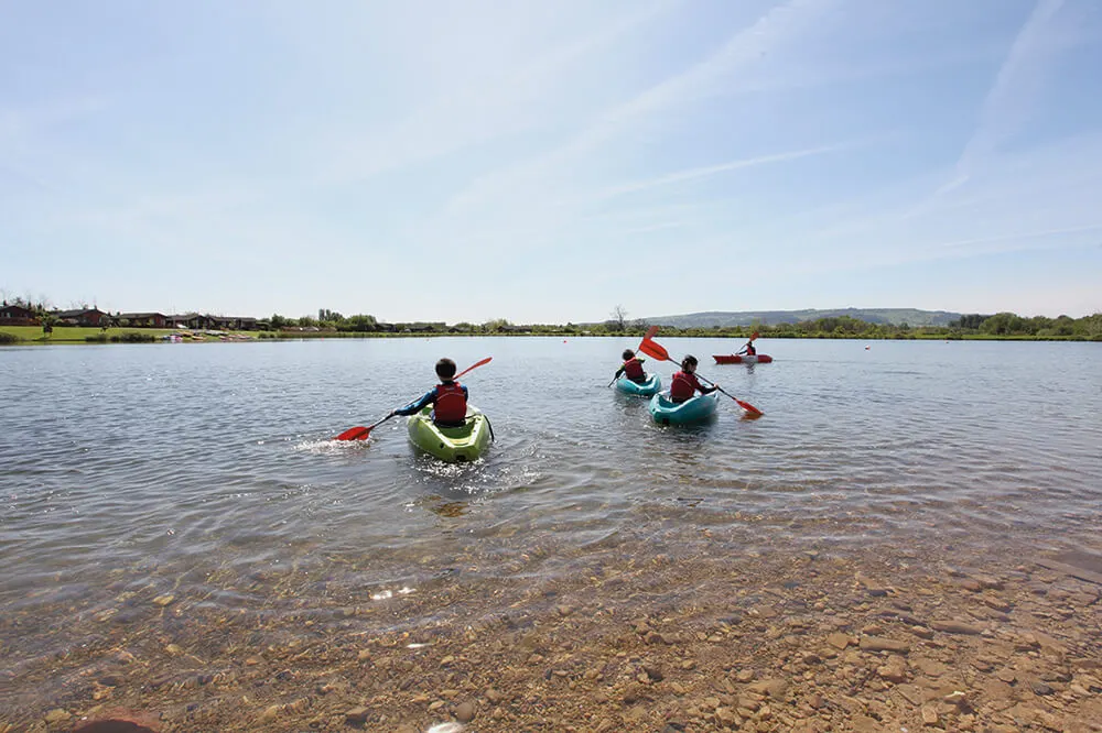People paddling kayaks on a calm lake with trees and hills visible along the shoreline.