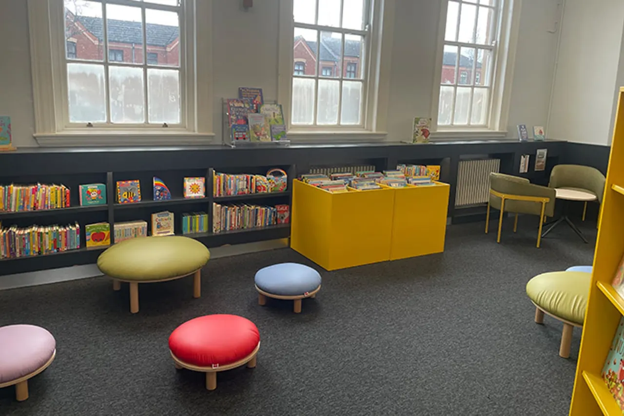 A modern children's library room with colourful cushioned seating stools, yellow chairs, black shelving filled with children's books, and large windows overlooking brick buildings.