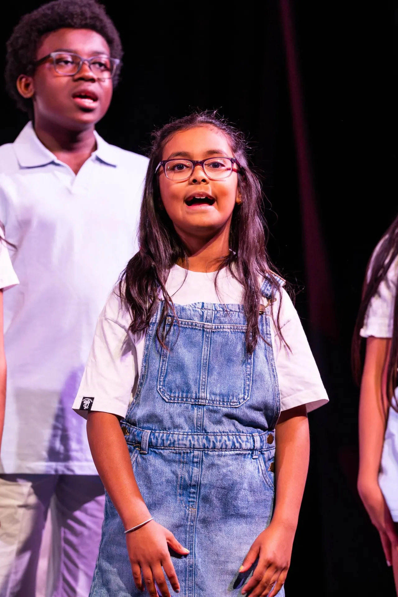 Children performing on stage under pink and purple stage lighting, singing together.
