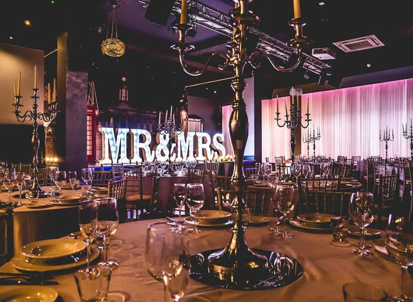 Elegant wedding reception hall with round tables set with place settings, gold candelabras, and glassware beneath purple lighting and an illuminated 'MR & MRS' sign.