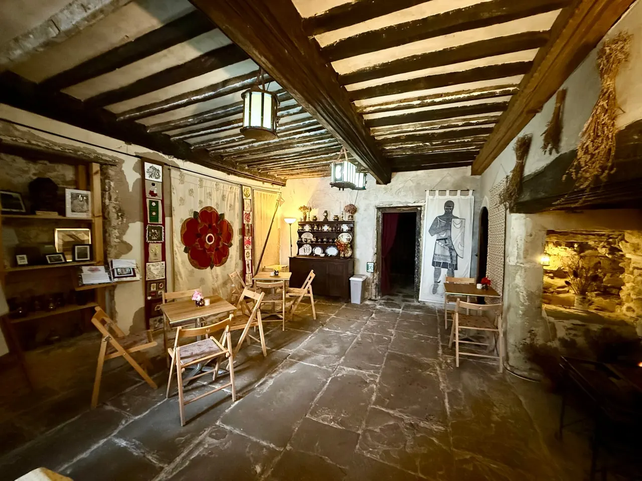 Interior of a period room with exposed wooden beams, stone flooring, wooden chairs and tables, decorated with historical artefacts, artwork and a large red flower ornament.