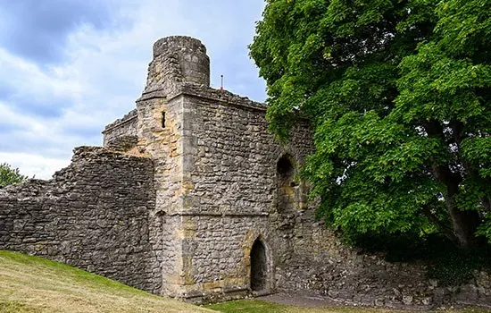 Medieval stone castle tower with crenellations and arched doorways, partially ivy-clad, set against cloudy sky.