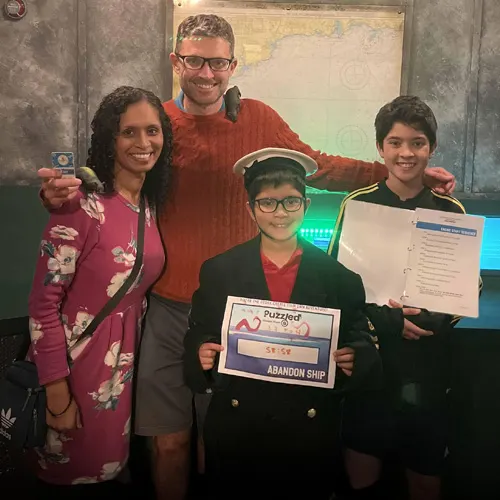 Four people of varying ages pose together indoors holding certificates and a puzzle game card after completing an escape room challenge.