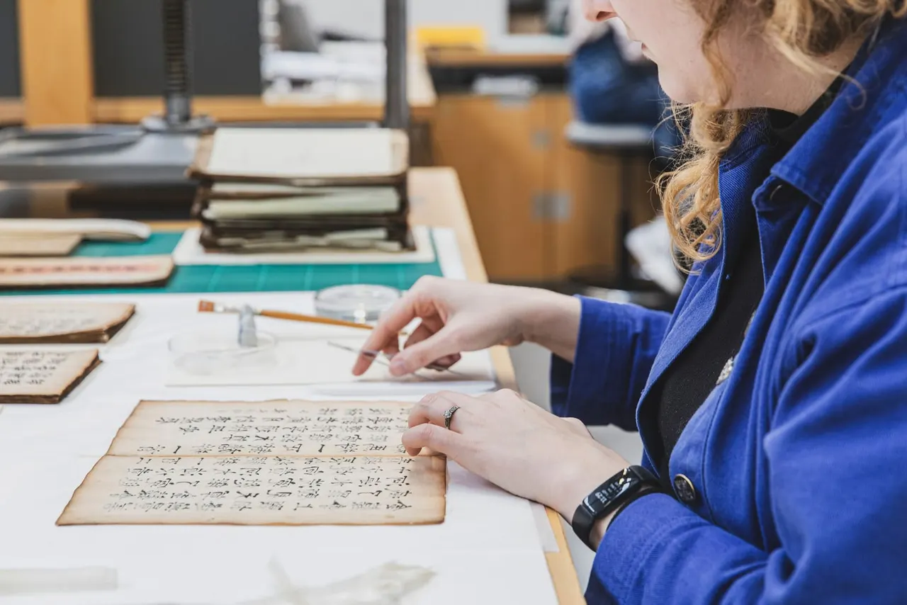 Woman examining a handwritten manuscript document on a table with other historical papers and archival materials.